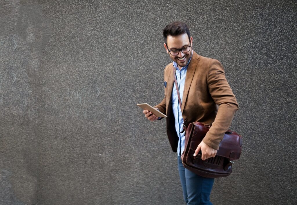 Ein lächelnder Mann mit Brille und braunem Blazer hält in der einen Hand ein Tablet und in der anderen eine Ledertasche, während er vor einer strukturierten grauen Wand steht.