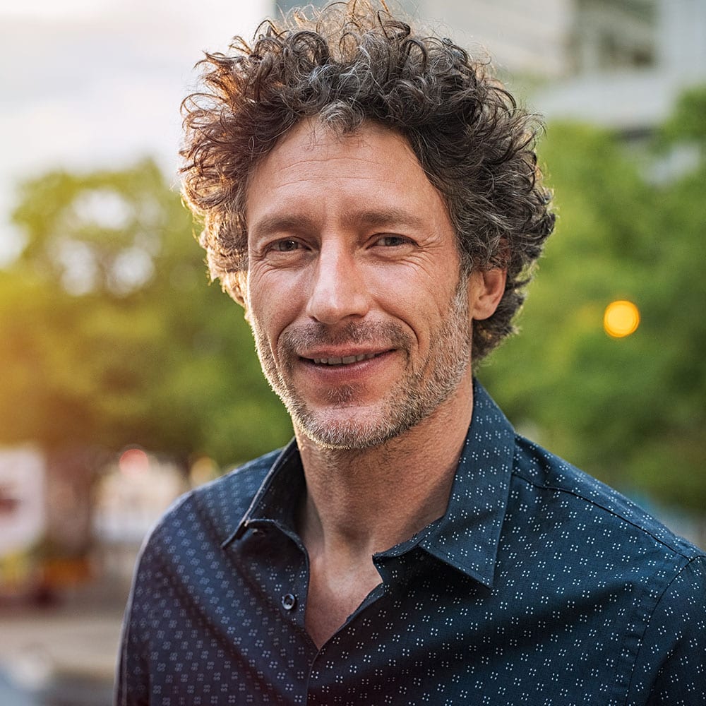 Ein Mann mit lockigem braunem Haar und hellem Bart lächelt im Freien und trägt ein dunkel gemustertes Hemd. Im Hintergrund sind Bäume und unscharfe Stadtgebäude zu sehen, wobei warmes Sonnenlicht die Szene beleuchtet.