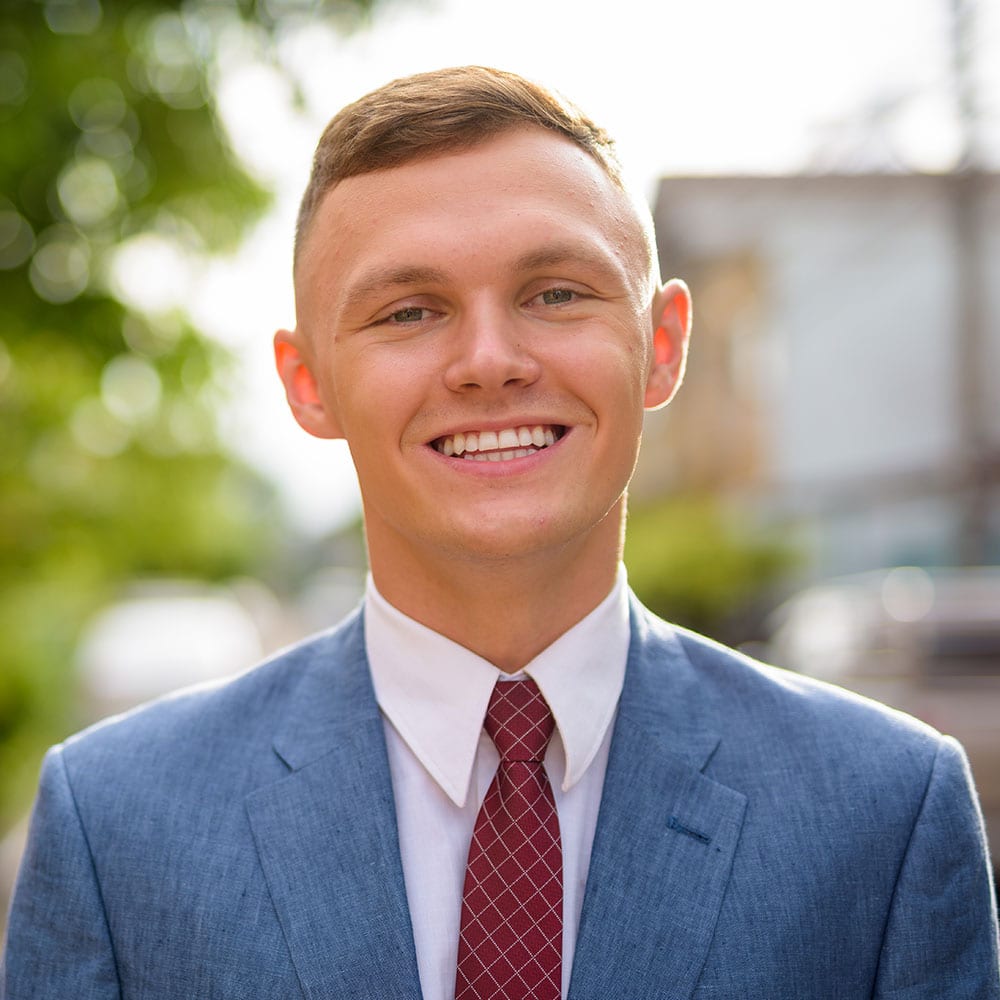 Ein junger Mann in blauem Anzug, weißem Hemd und roter Krawatte lächelt im Freien, vor einem unscharfen Hintergrund aus Grünflächen und Gebäuden.